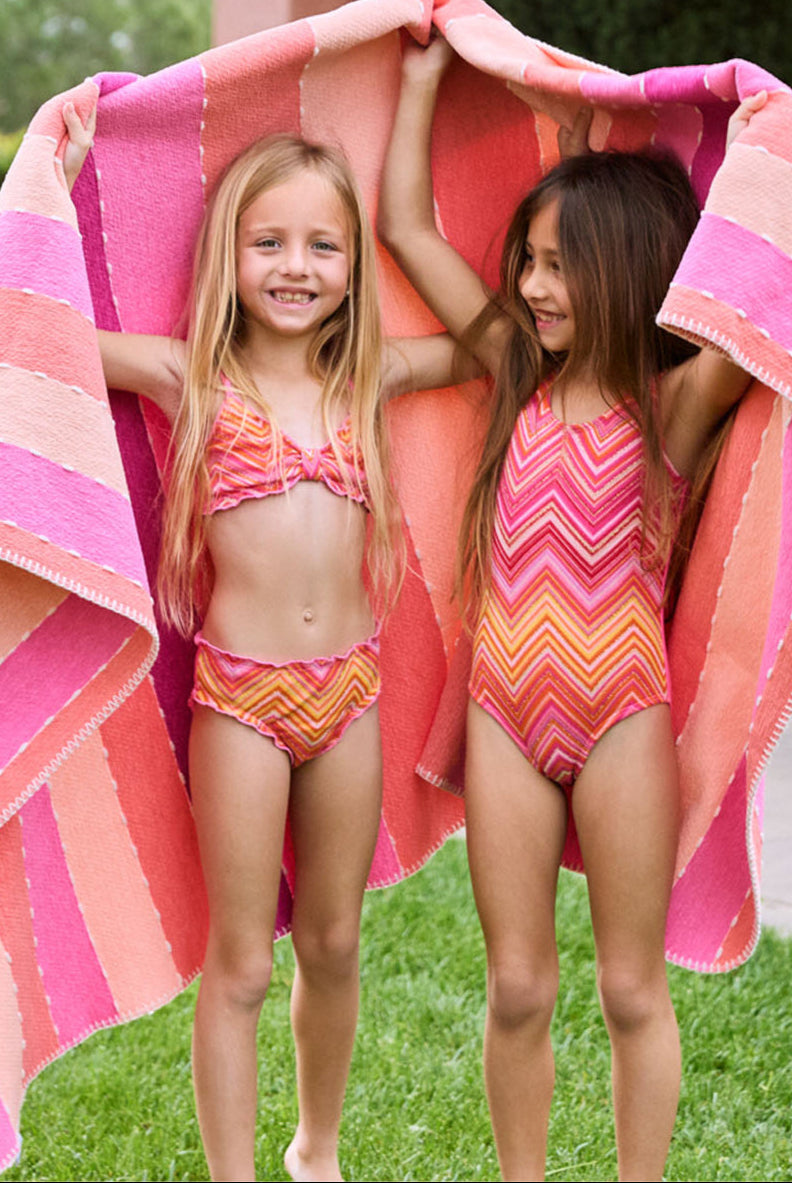 A blonde girl wearing a pink and orange striped bikini and a brunette girl wearing an orange and pink striped one piece. Both girls are standing on the grass outside.