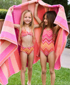 A blonde girl wearing a pink and orange striped bikini and a brunette girl wearing an orange and pink striped one piece. Both girls are standing on the grass outside.