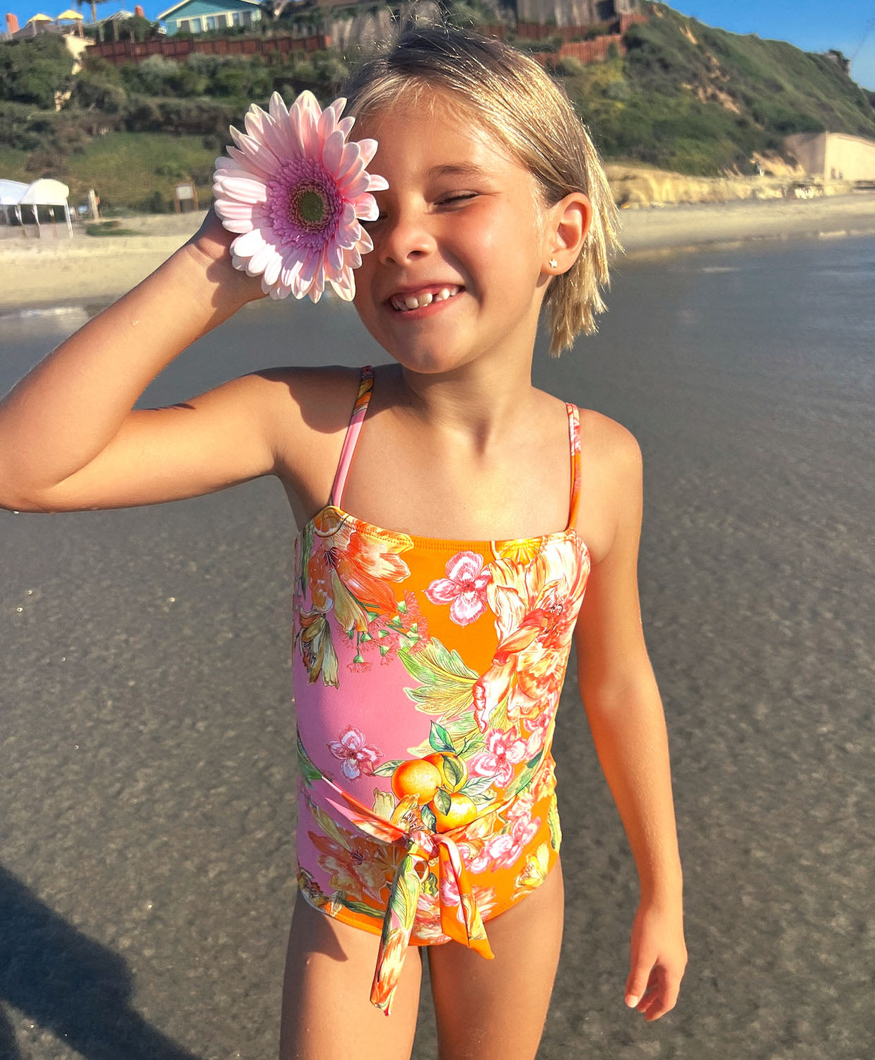 A young girl wearing a floral one piece swim suit standing on a beach near an ocean.