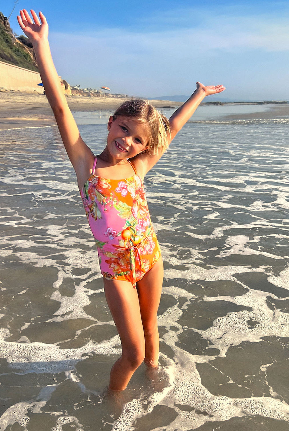 A young girl wearing a floral one piece swim suit standing on a beach near an ocean.