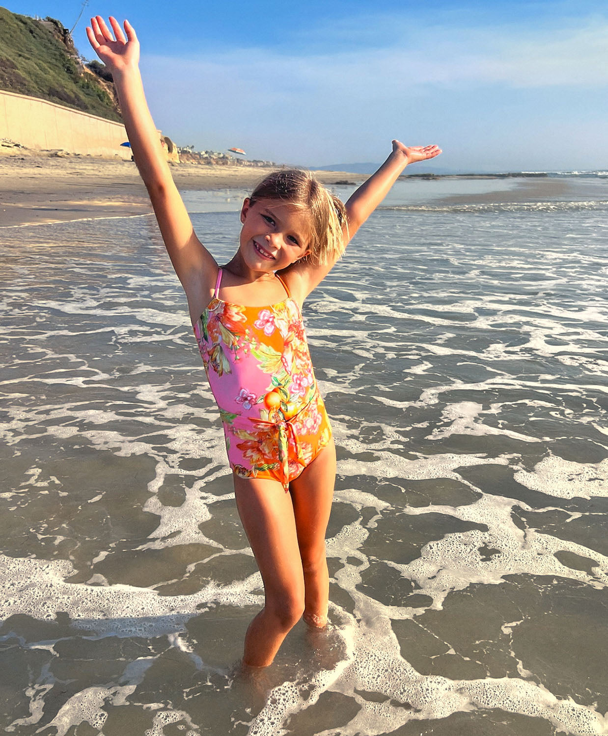 A young girl wearing a floral one piece swim suit standing on a beach near an ocean.