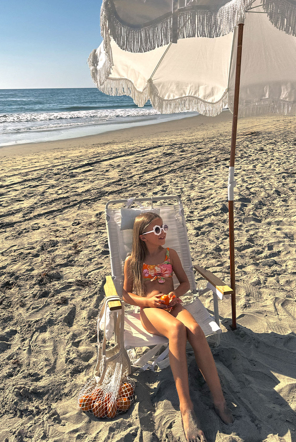 A young girl wearing a pink and orange bikini sitting on a chair under an umbrella.