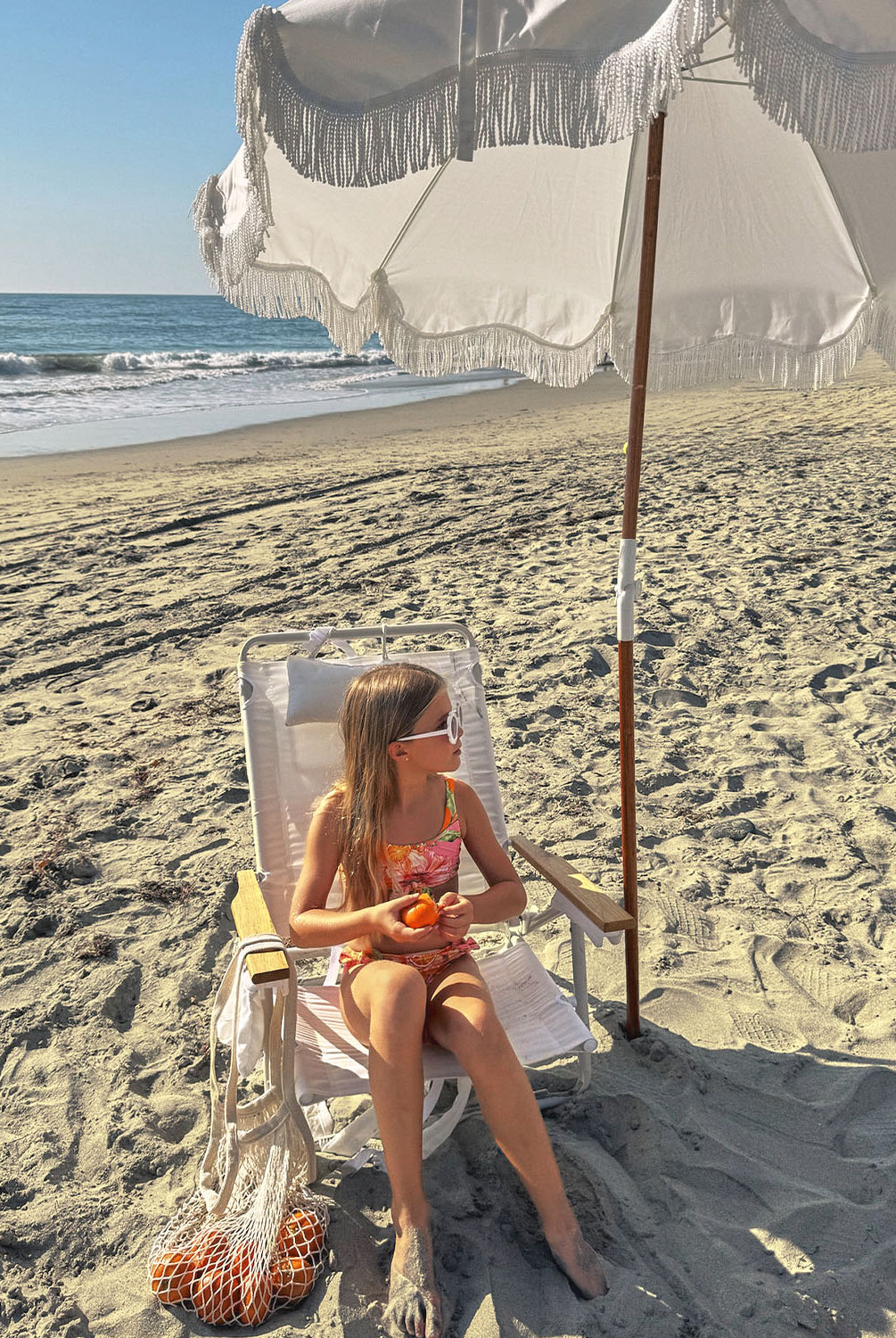 A young girl wearing a pink and orange bikini sitting on a chair under an umbrella.