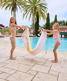 A blonde girl wearing a light pink one piece suit and a brunette girl wearing a multi colored bikini. Both girls are standing near a pool.