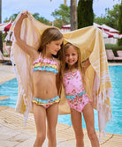 To girls wearing floral swimsuits holding up a beach towel near a pool.
