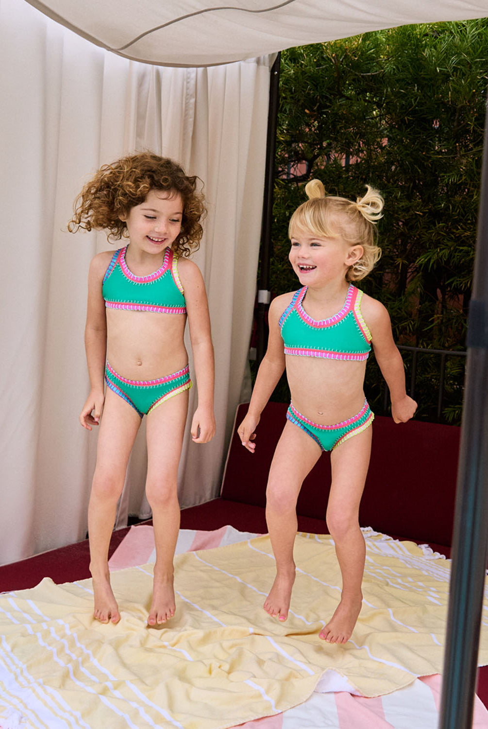 Two girls wearing green bikinis with colorful embroidery standing on a beach towel.