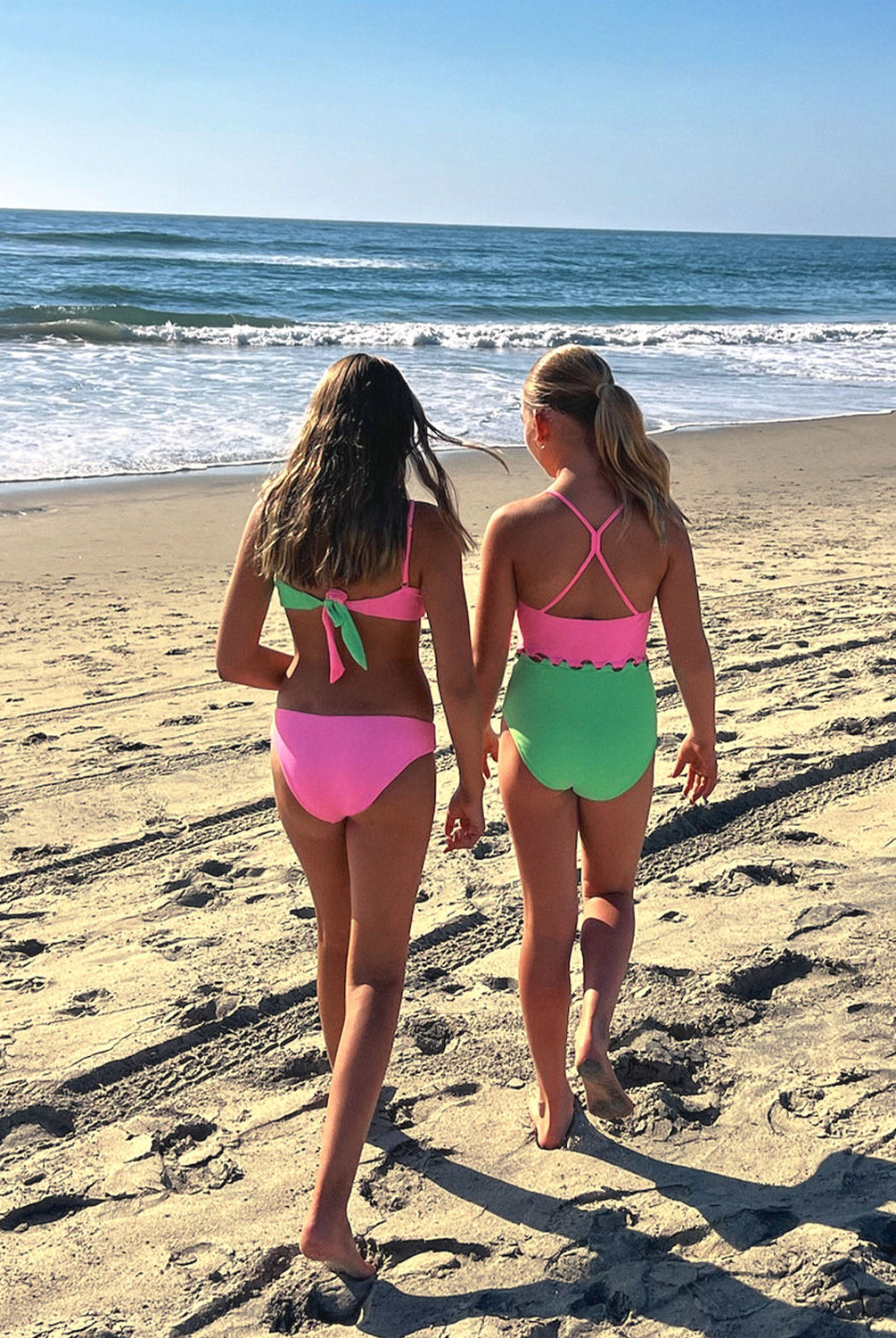 Two young girls standing on a beach near an ocean. One girl is wearing a pink and green bikini and the other girl is wearing a pink and green one piece.