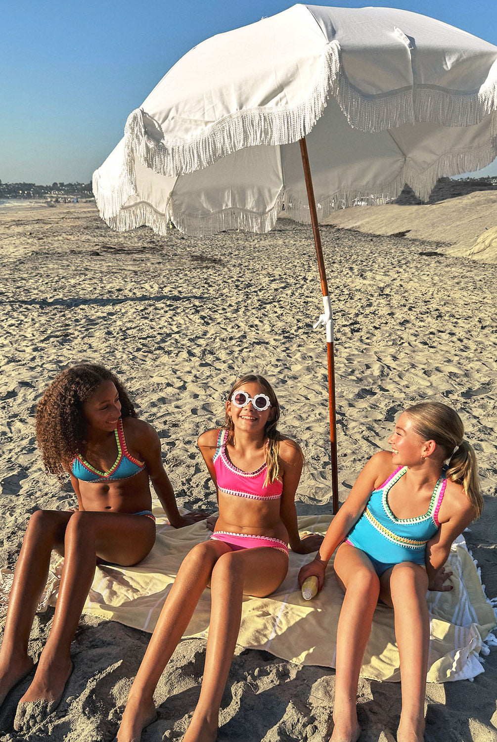 Three young girls sitting on a beach under an umbrella. One girl is wearing a turquoise bikini, another girl is wearing a hot pink bikini, and another girl is wearing a turquoise one piece.