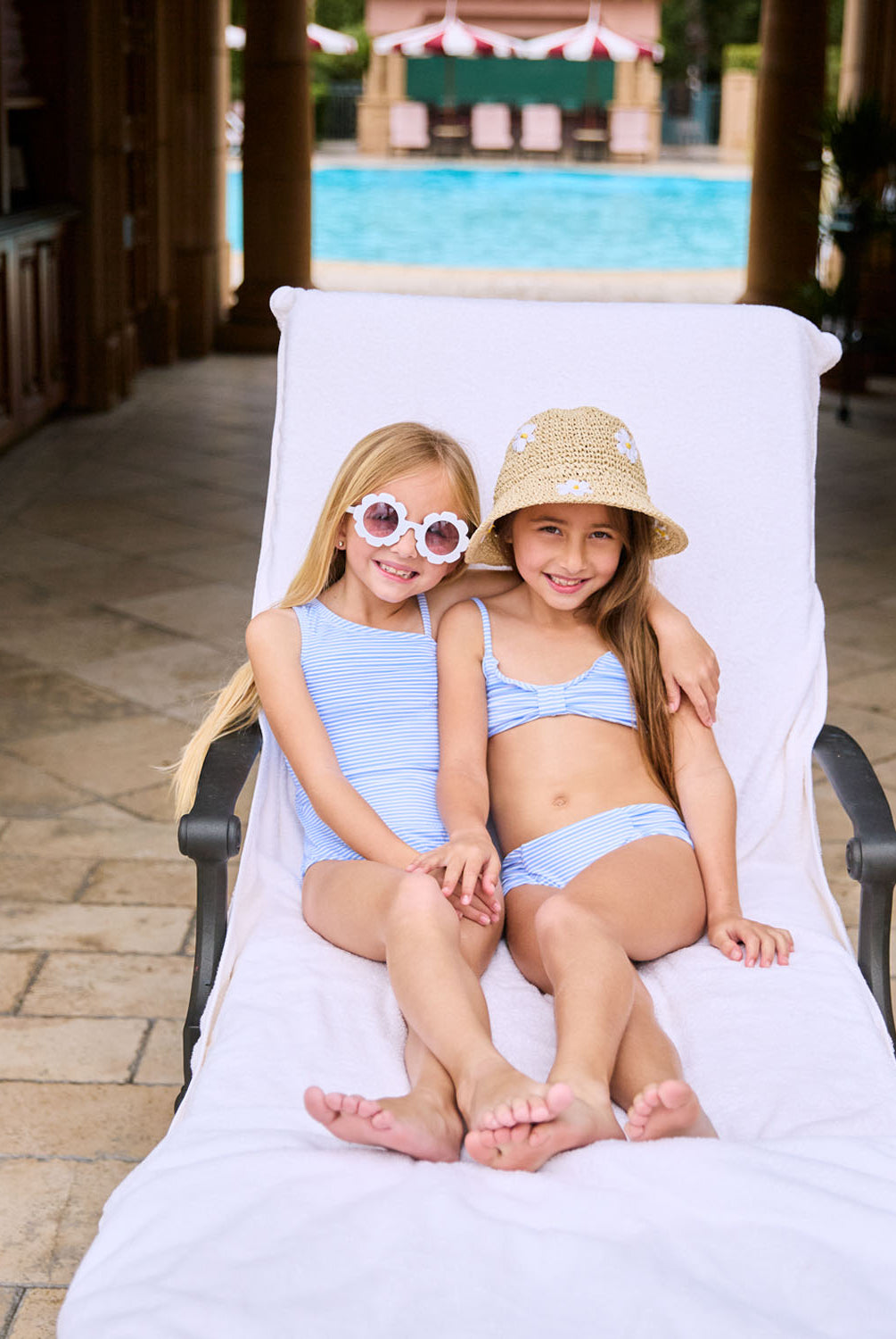 A blonde girl wearing a blue and white striped one piece swimsuit. A brunette girl wearing a blue and white striped bikini. Both girls are lounging on a lawn chair.