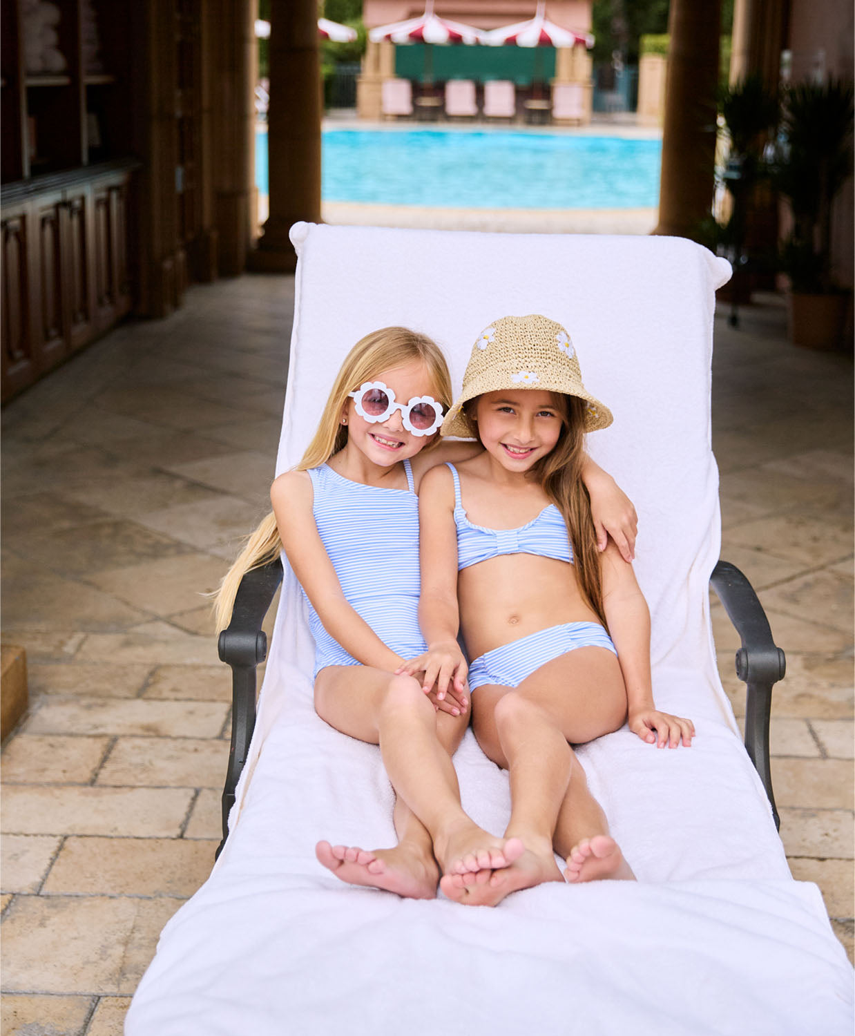 A blonde girl wearing a blue and white striped one piece swimsuit. A brunette girl wearing a blue and white striped bikini. Both girls are lounging on a lawn chair.