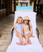 A blonde girl wearing a blue and white striped one piece swimsuit. A brunette girl wearing a blue and white striped bikini. Both girls are lounging on a lawn chair.