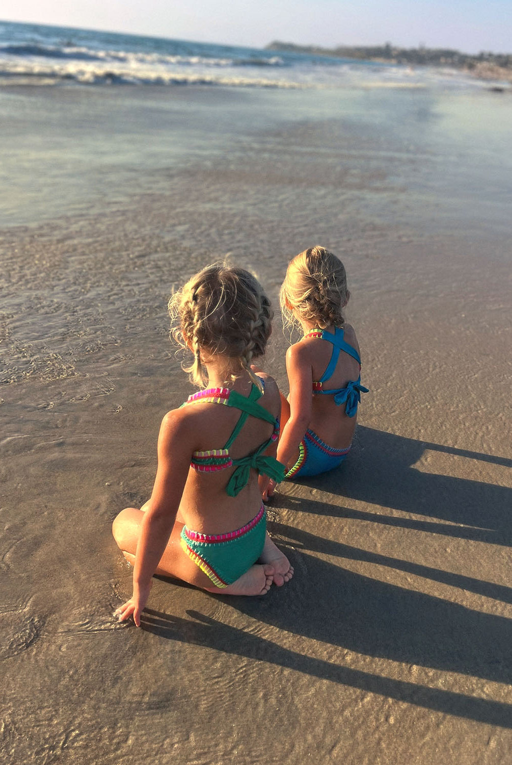 Two girls sitting on a beach near the ocean. One girl is wearing a green embroidered bikini and the other girl is wearing a turquoise embroidered bikini.