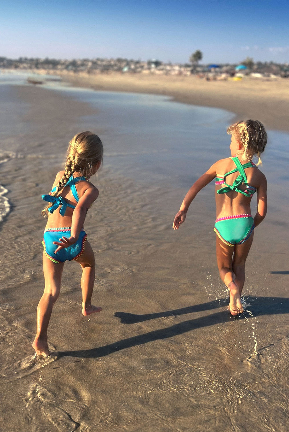 Two girls walking on a beach near the ocean. One girl is wearing a green embroidered bikini and the other girl is wearing a turquoise embroidered bikini.