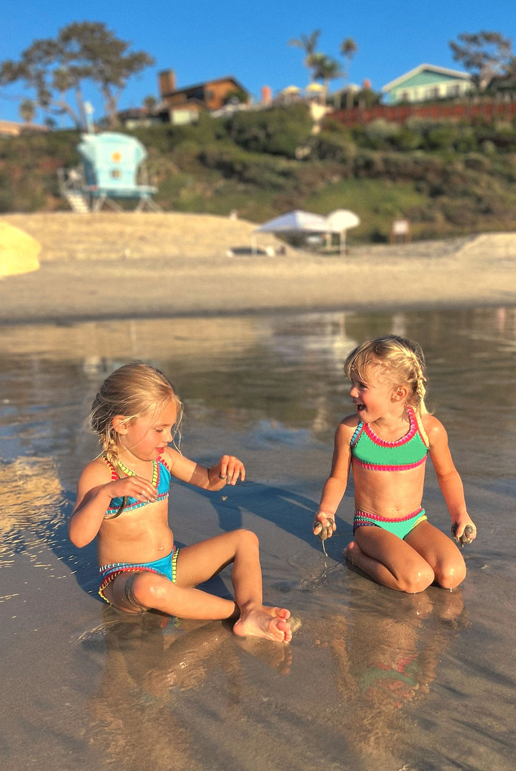 Two girls are sitting on a beach. One girl is wearing a turquoise bikini and the other girl is wearing a green bikini.