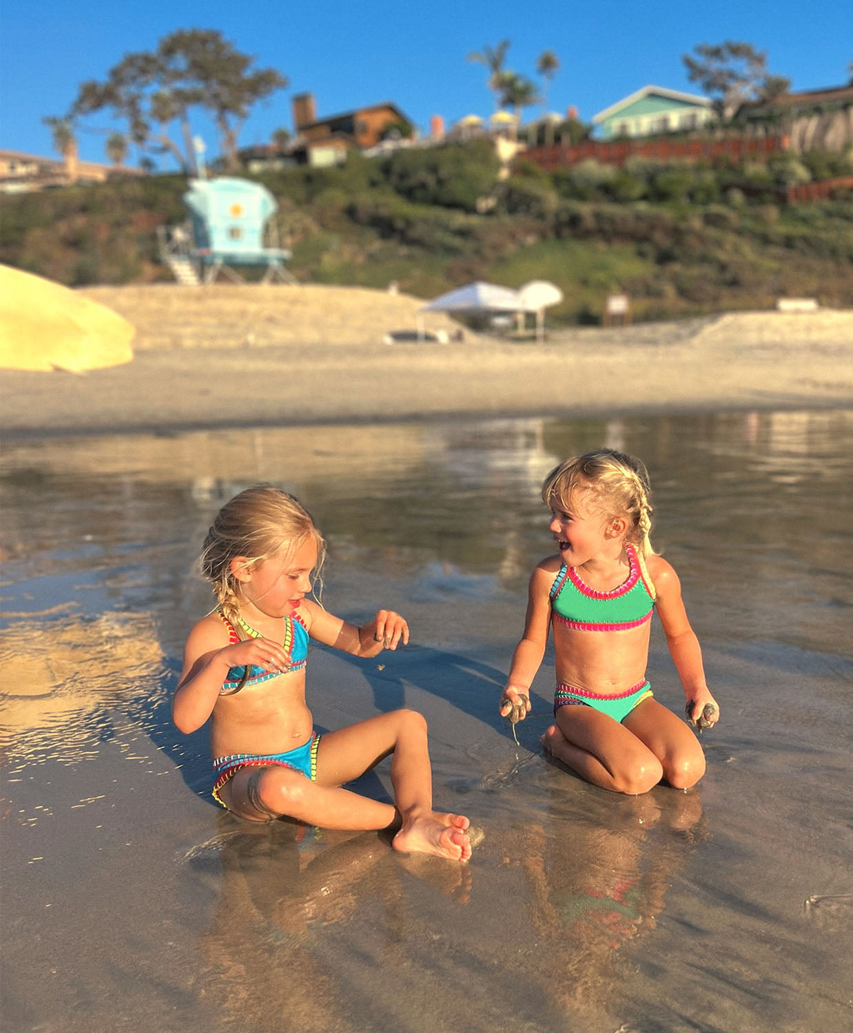 Two girls are sitting on a beach. One girl is wearing a turquoise bikini and the other girl is wearing a green bikini.