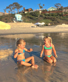 Two girls are sitting on a beach. One girl is wearing a turquoise bikini and the other girl is wearing a green bikini.