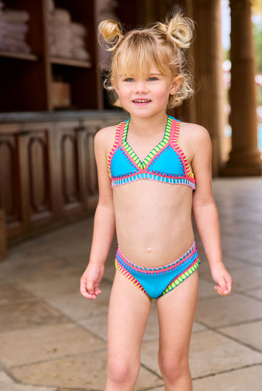 A blonde girl wearing a turquoise bikini with colorful embroidery standing near a pool.