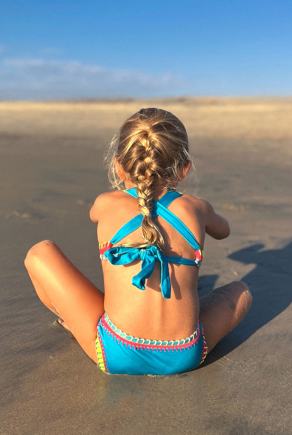 A girl wearing a turquoise bikini with embroidered trim sitting on a beach.