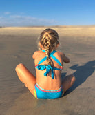 A girl wearing a turquoise bikini with embroidered trim sitting on a beach.