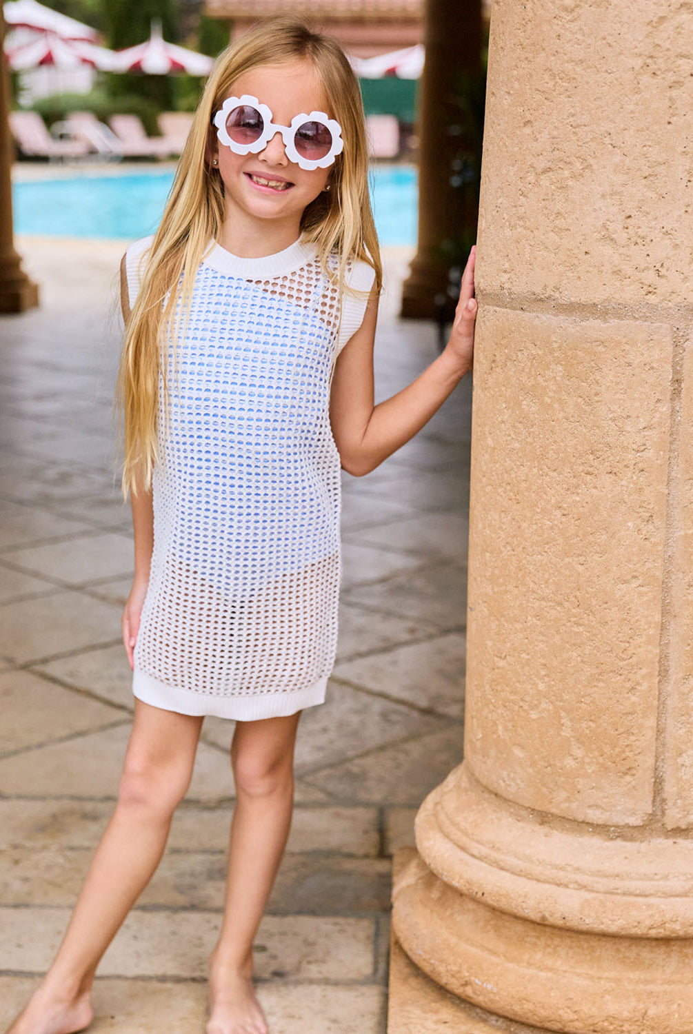 A blonde girl wearing a white knit cover up dress standing near a pool.