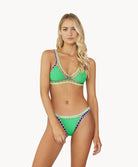 A blonde woman wearing a green bikini stands in front of a white wall. 