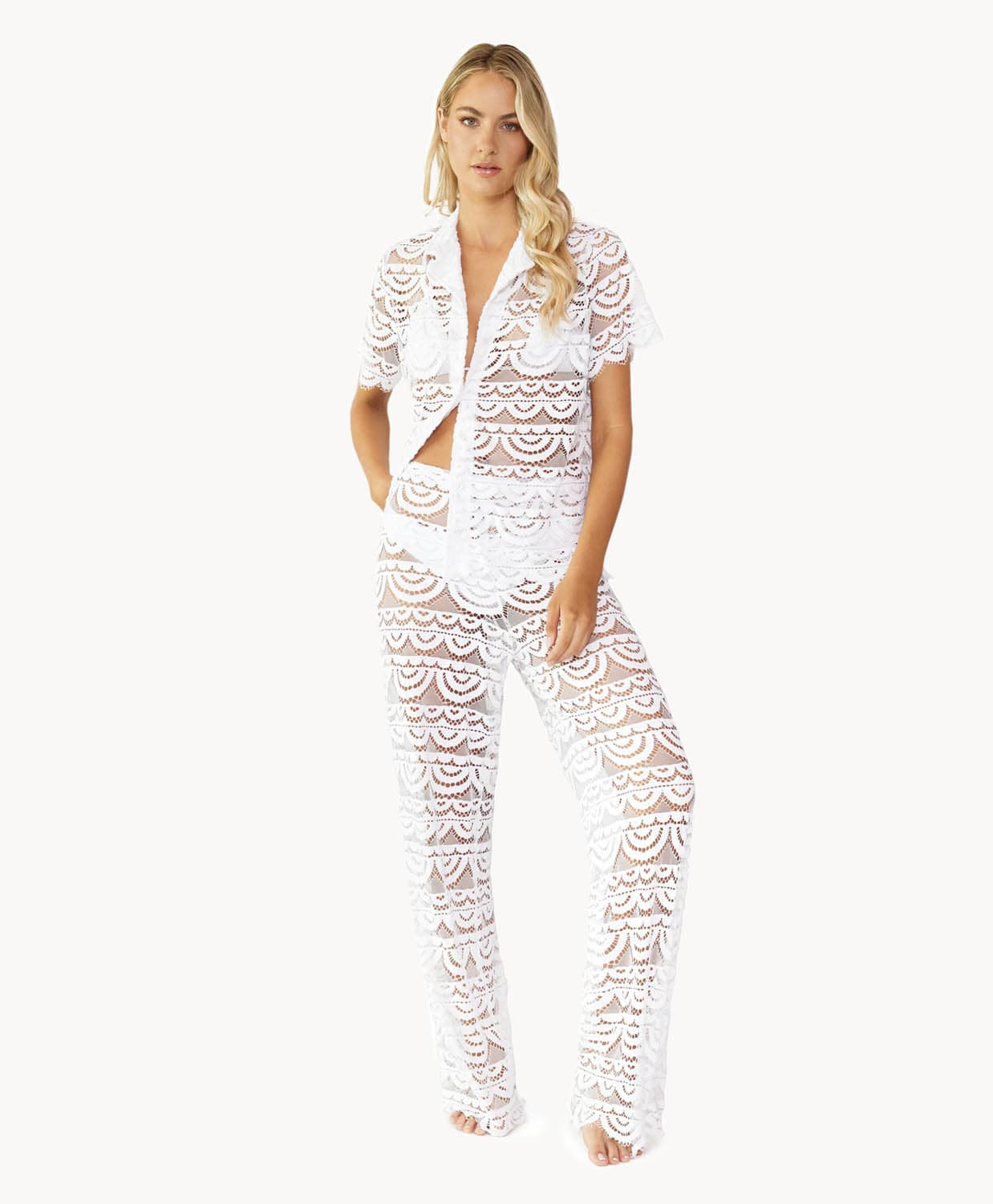 Woman wearing a matching white lace button shirt and pant against white wall.