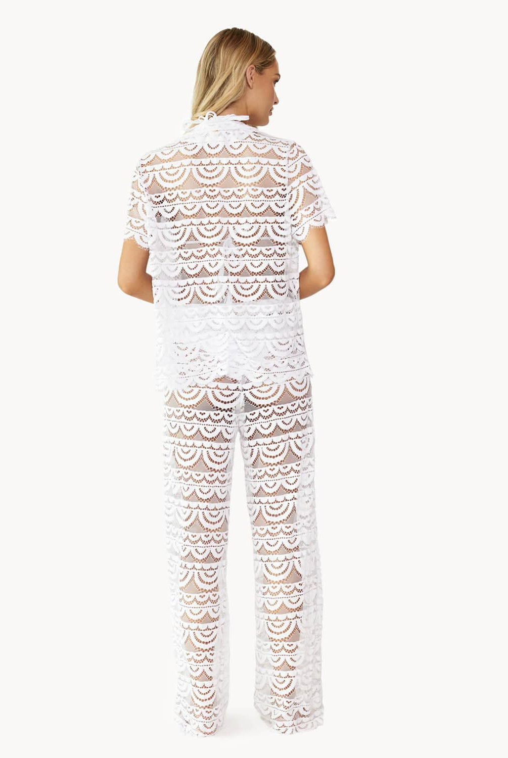 Back view of woman wearing a matching white lace button shirt and pant against white wall.
