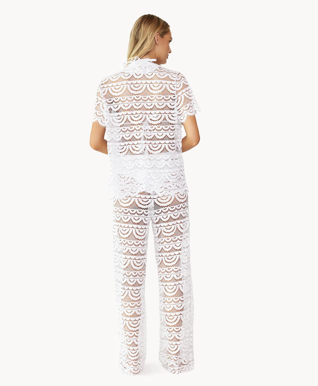 Back view of woman wearing a matching white lace button shirt and pant against white wall.