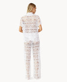 Back view of woman wearing a matching white lace button shirt and pant against white wall.