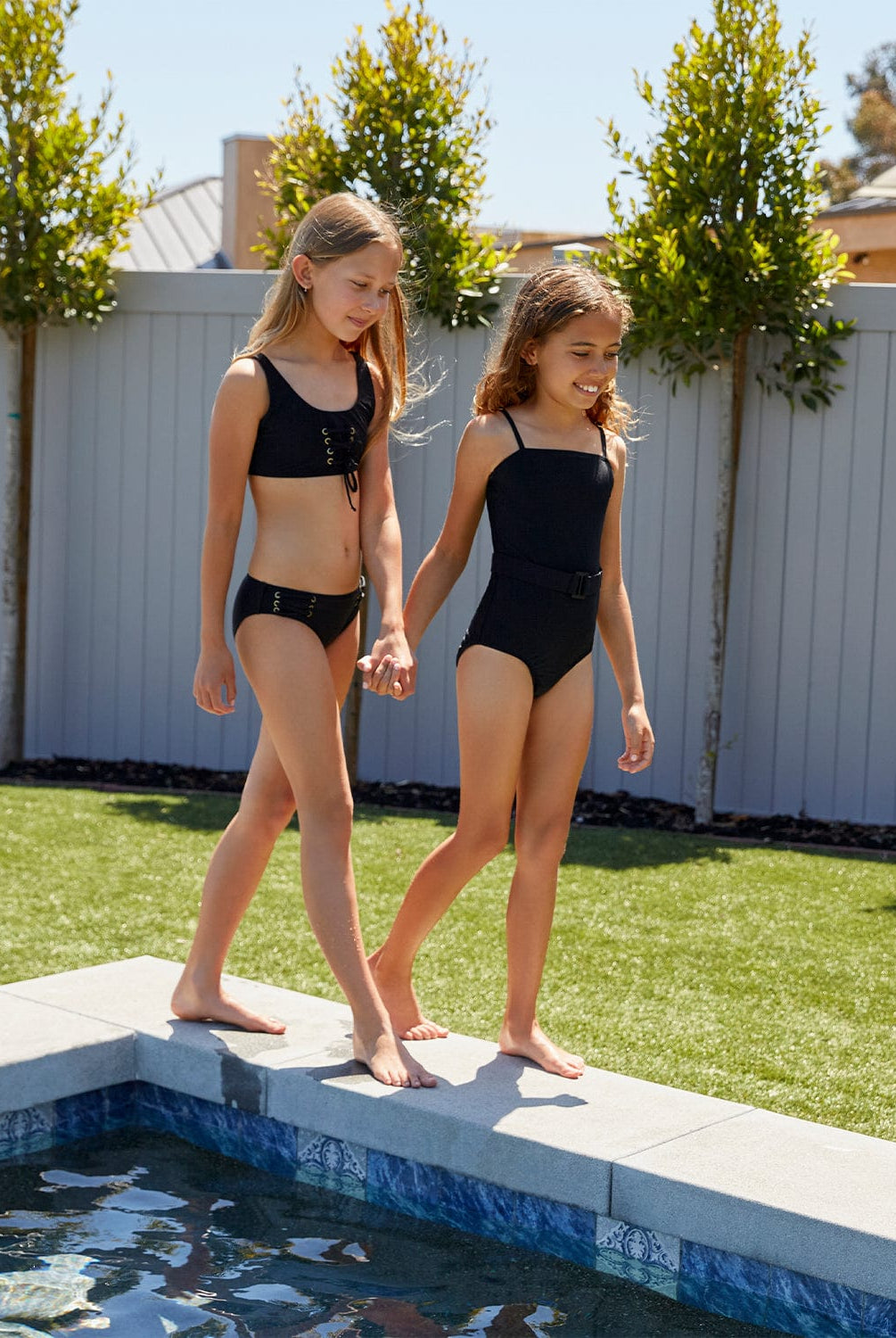 Blonde girl in black bikini and a brunette girl in black one piece walking by a pool.