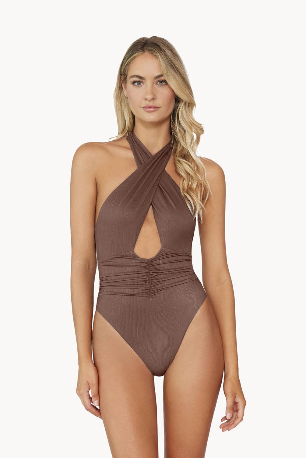 A blonde woman wearing a brown one piece bathing suit stands in front of a white wall. 