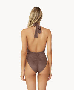 A blonde woman wearing a brown one piece bathing suit facing a white wall. 