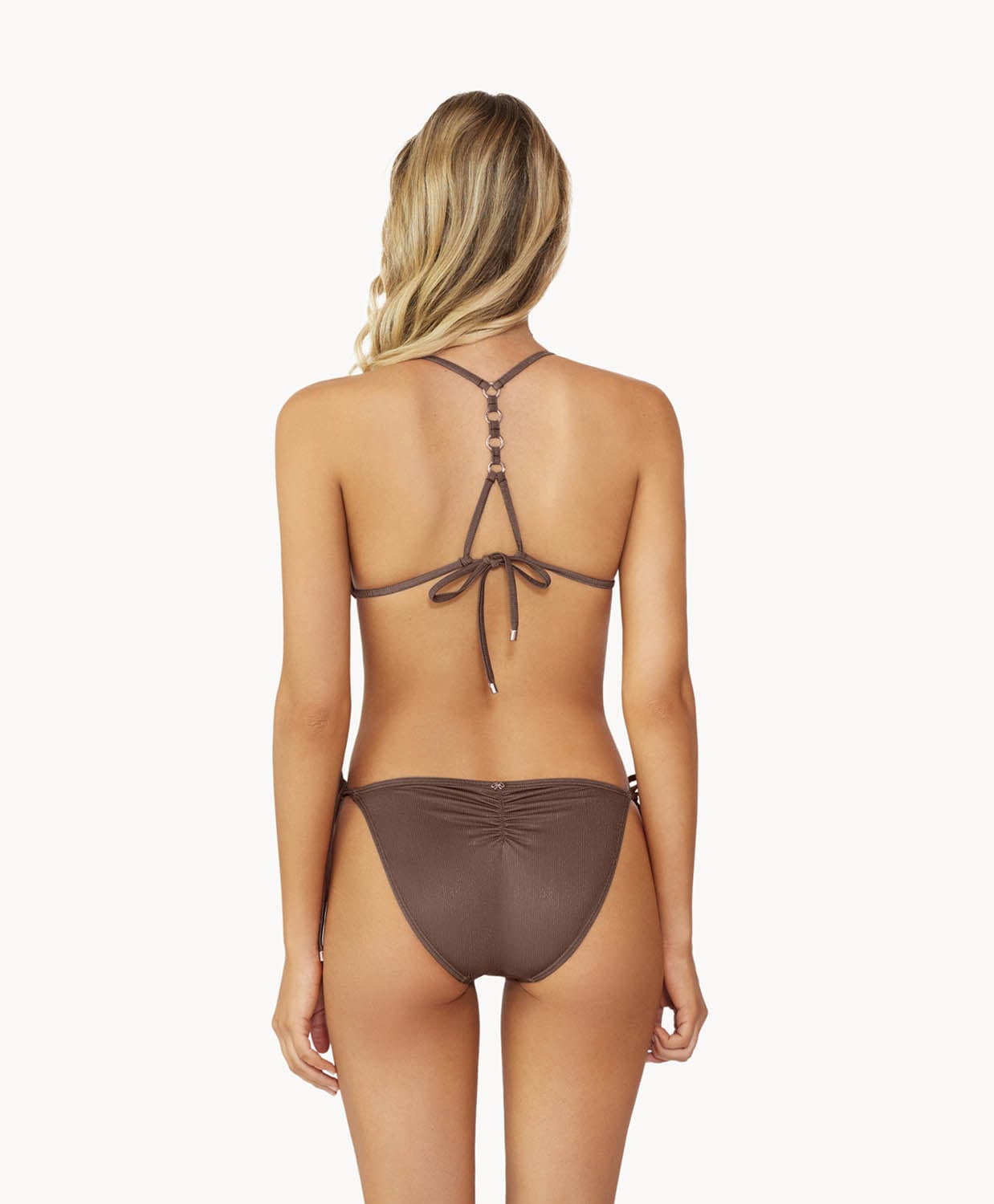 A blonde woman wearing a brown bikini facing towards a white wall.