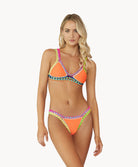 A blonde woman wearing an orange bikini stands in front of a white wall.