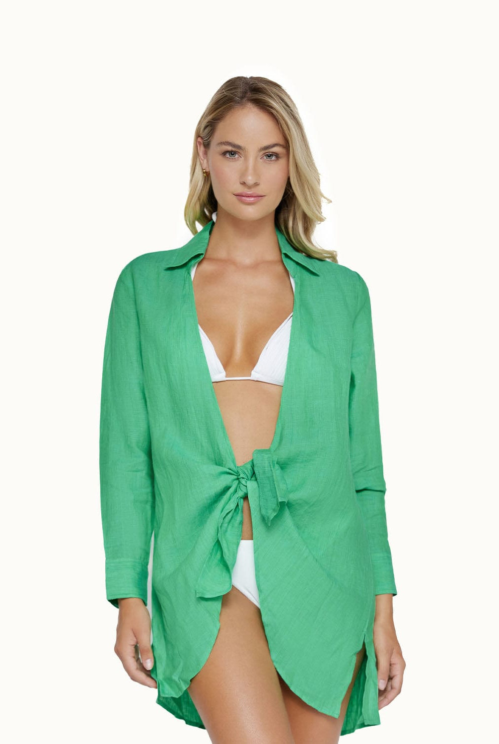 A blonde woman wearing a white bikini and a green long sleeve cover up against a white wall. 