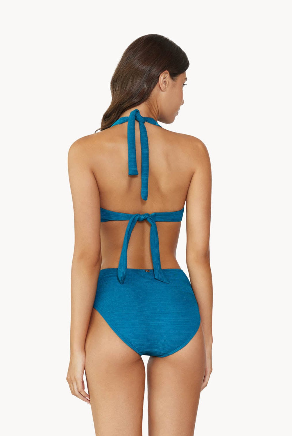 Brunette woman wearing a textured turquoise halter bikini with gold details facing backwards towards a white wall.