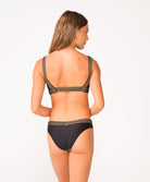 A woman wearing a black bikini with gold details facing towards a white wall.
