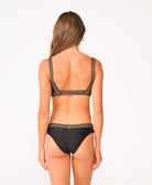 A woman wearing a black bikini with gold details facing towards a white wall.
