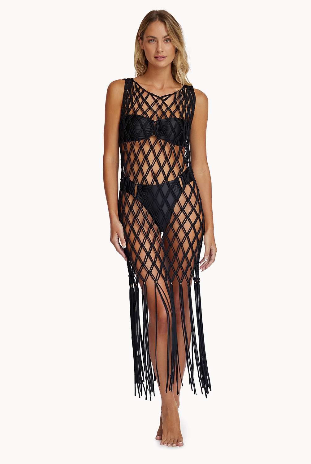 A woman wearing a black crochet cover up with fringe standing against a white wall.