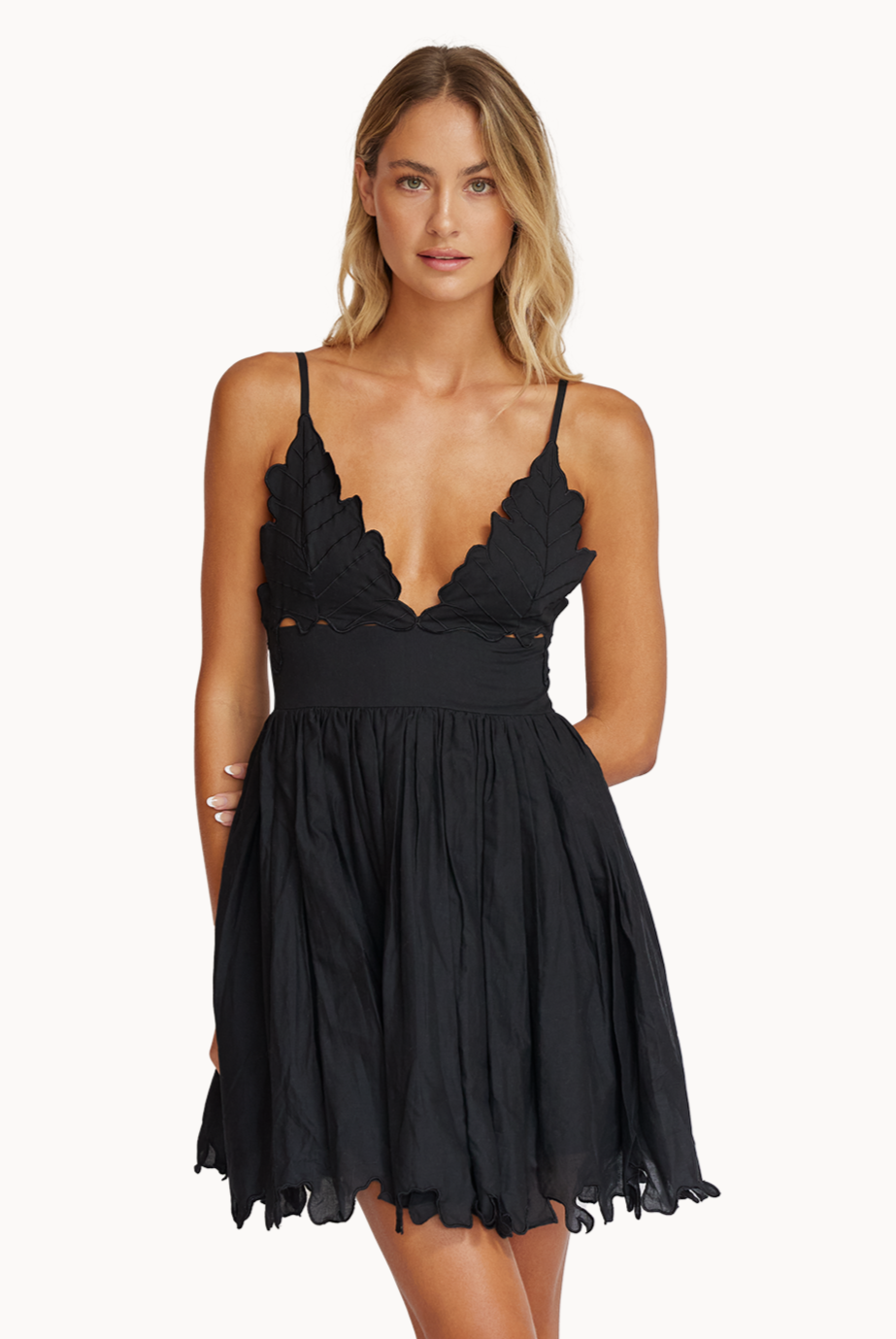 A blonde woman wearing a short black dress standing against a white wall.