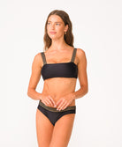A woman wearing a black bikini with gold details standing against a white wall.