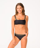 A woman wearing a black bikini with gold details standing against a white wall.