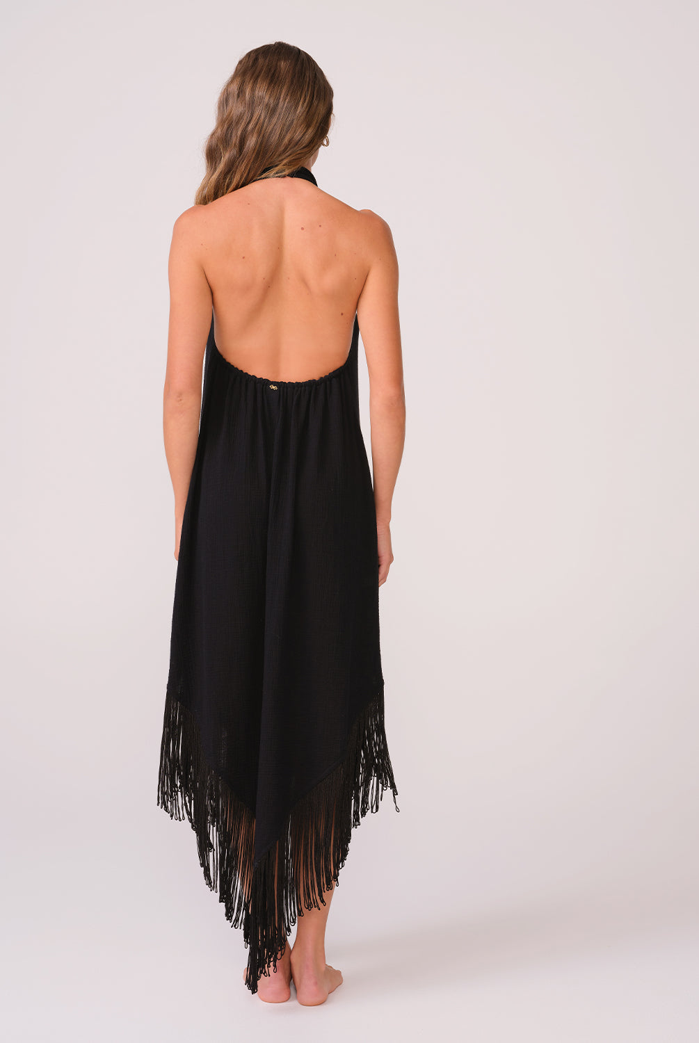 A woman wearing a long black fringe dress facing towards a white wall.