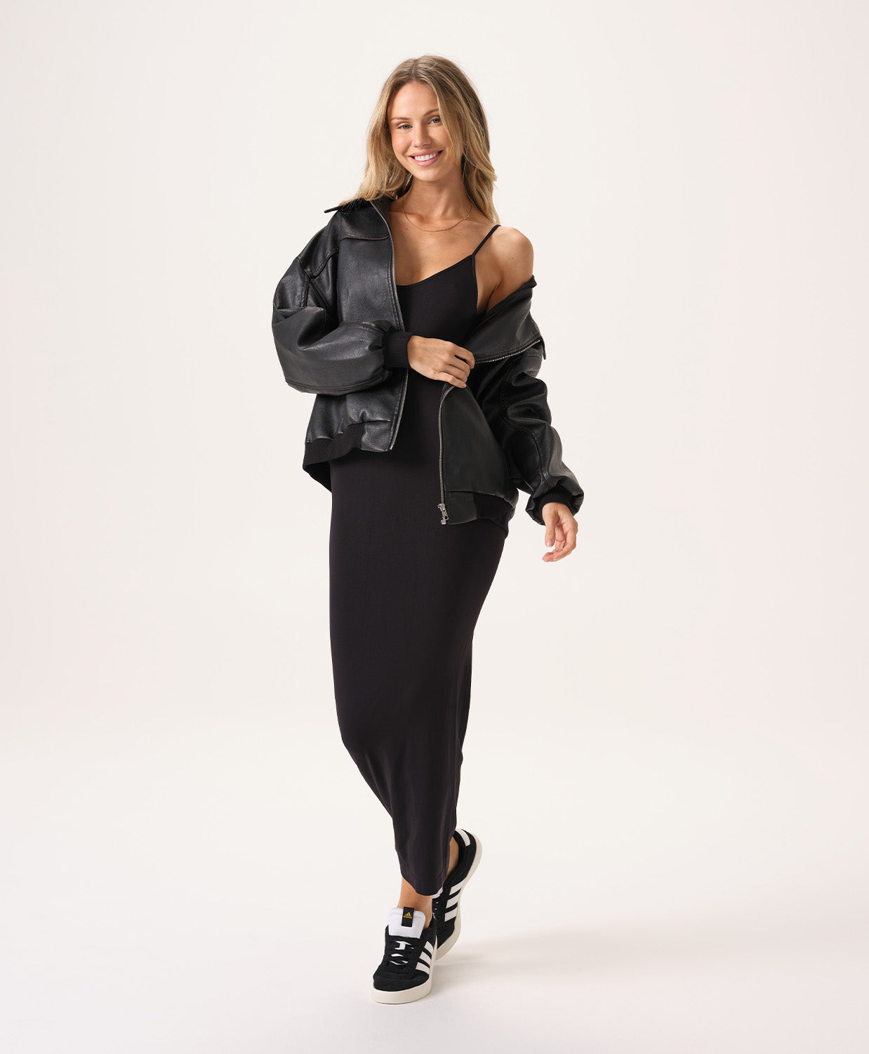 A woman wearing a long black slip dress and a black leather jacket standing against a white wall.