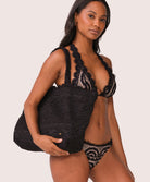 A woman holding a black lace tote with scalloped trim against a white wall.