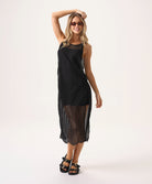 A woman wearing a short black slip dress under a black crochet dress standing against a white wall.