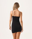 A woman wearing a short black slip dress facing towards a white wall.