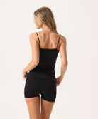 A woman wearing a black tank top and matching shorts facing towards a white wall.