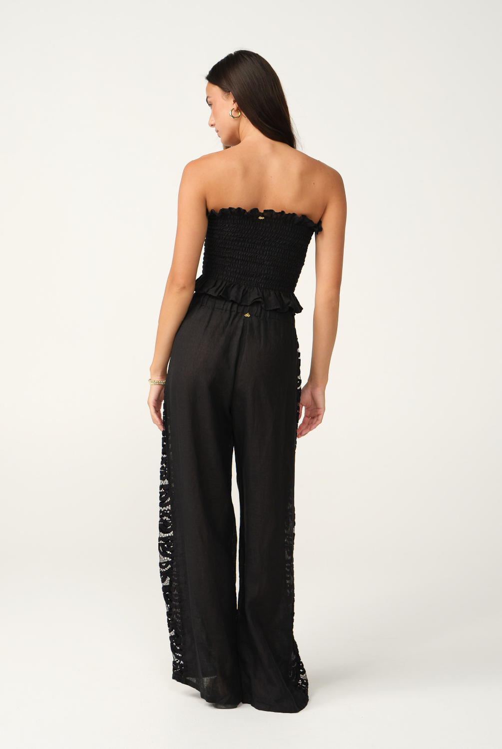 A woman modeling a black strapless cropped top and matching wide-leg pants with lace side panels, photographed against a clean off-white studio background.