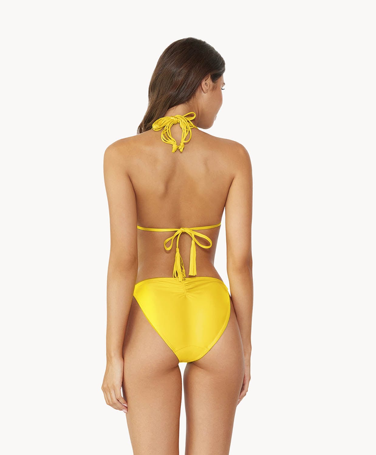 Brunette woman wearing a yellow macrame triangle shape bikini facing backwards towards a white wall.